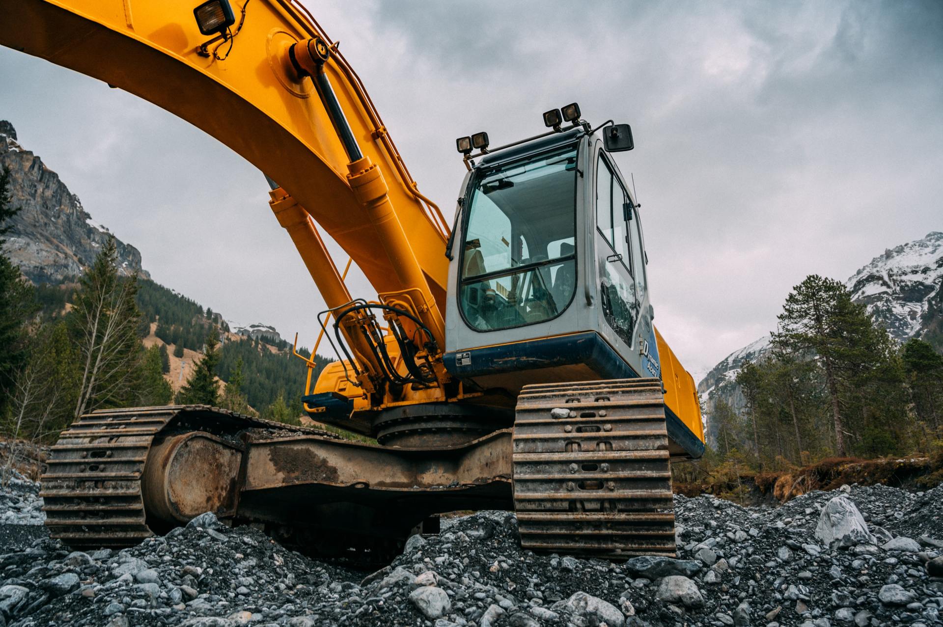 Yellow excavator on rocky terrain, mountains in background, cloudy sky.