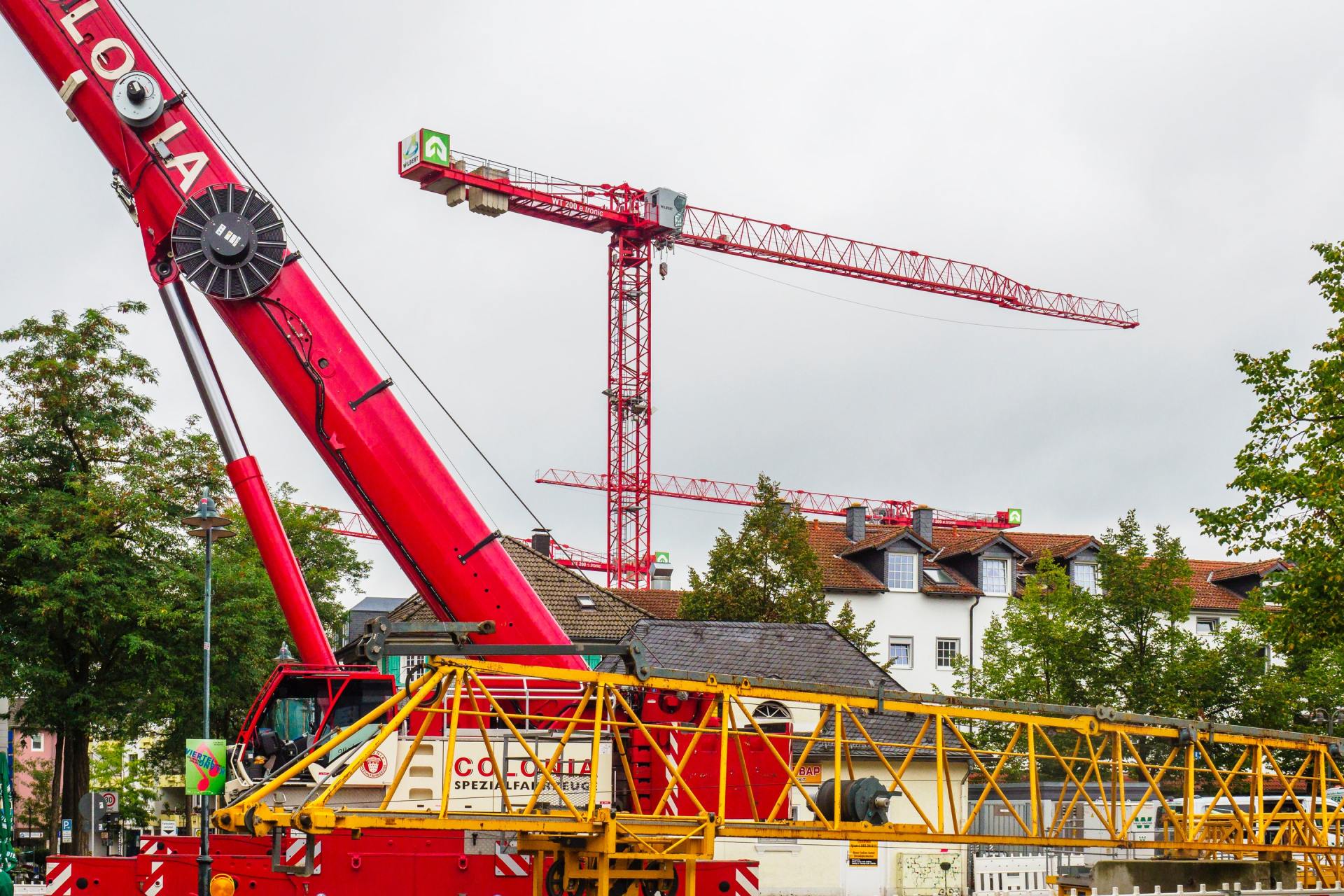 Red construction cranes on a cloudy day, with buildings and trees in the background.